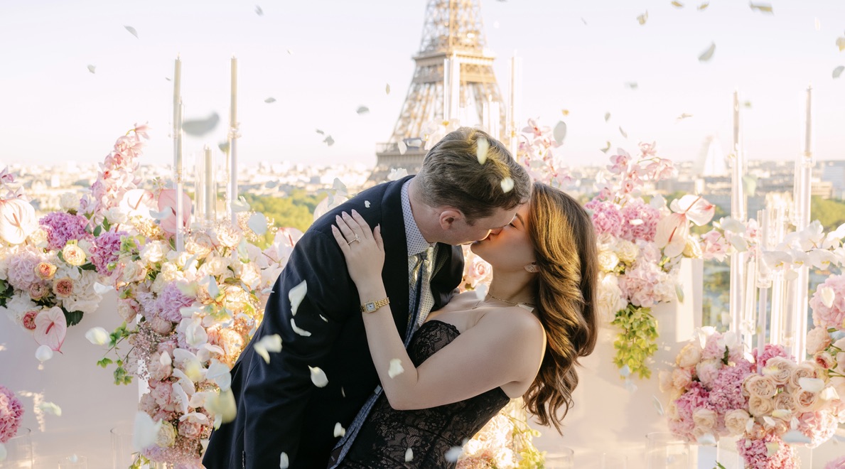 Couple kissing during a rooftop proposal in Paris with the Eiffel Tower in the background, surrounded by pink and white floral arrangements and candles