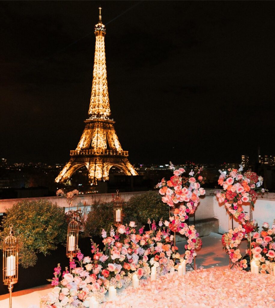 Romantic rooftop proposal setup in Paris at night with pink and coral flower arches, rose petals, candles, and gold lanterns facing the illuminated Eiffel Tower