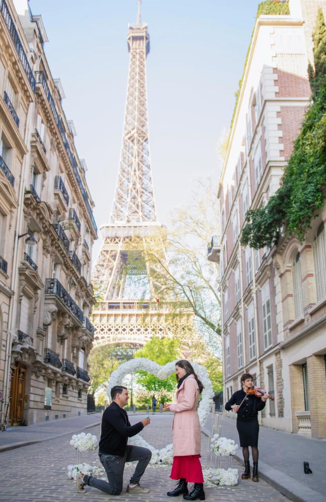 A person kneeling on one knee proposing to a woman on a cobblestone street in Paris, with the Eiffel Tower rising in the background. A heart-shaped floral arch and flower arrangements frame the couple, while a violinist performs nearby