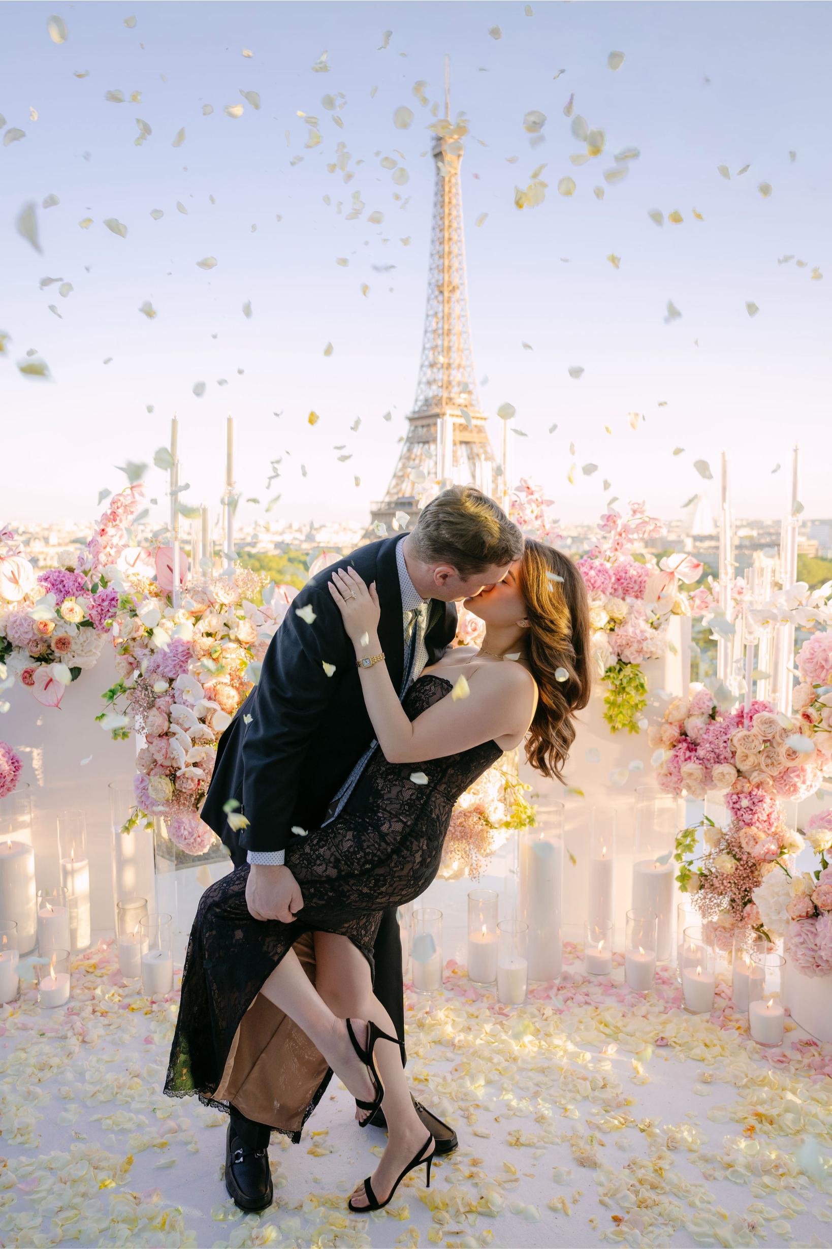 Couple kissing on the rooftop terrace of the Shangri-La Paris hotel, surrounded by pink and white floral arrangements, candles and rose petals falling from above, with a direct view of the Eiffel Tower in the background
