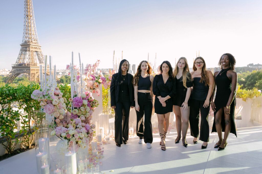 Un équipe de femme habillées en noir, pause en ligne au milieu d'un décor de fleurs et de chandeliers. avec la Tour Eiffel en fond