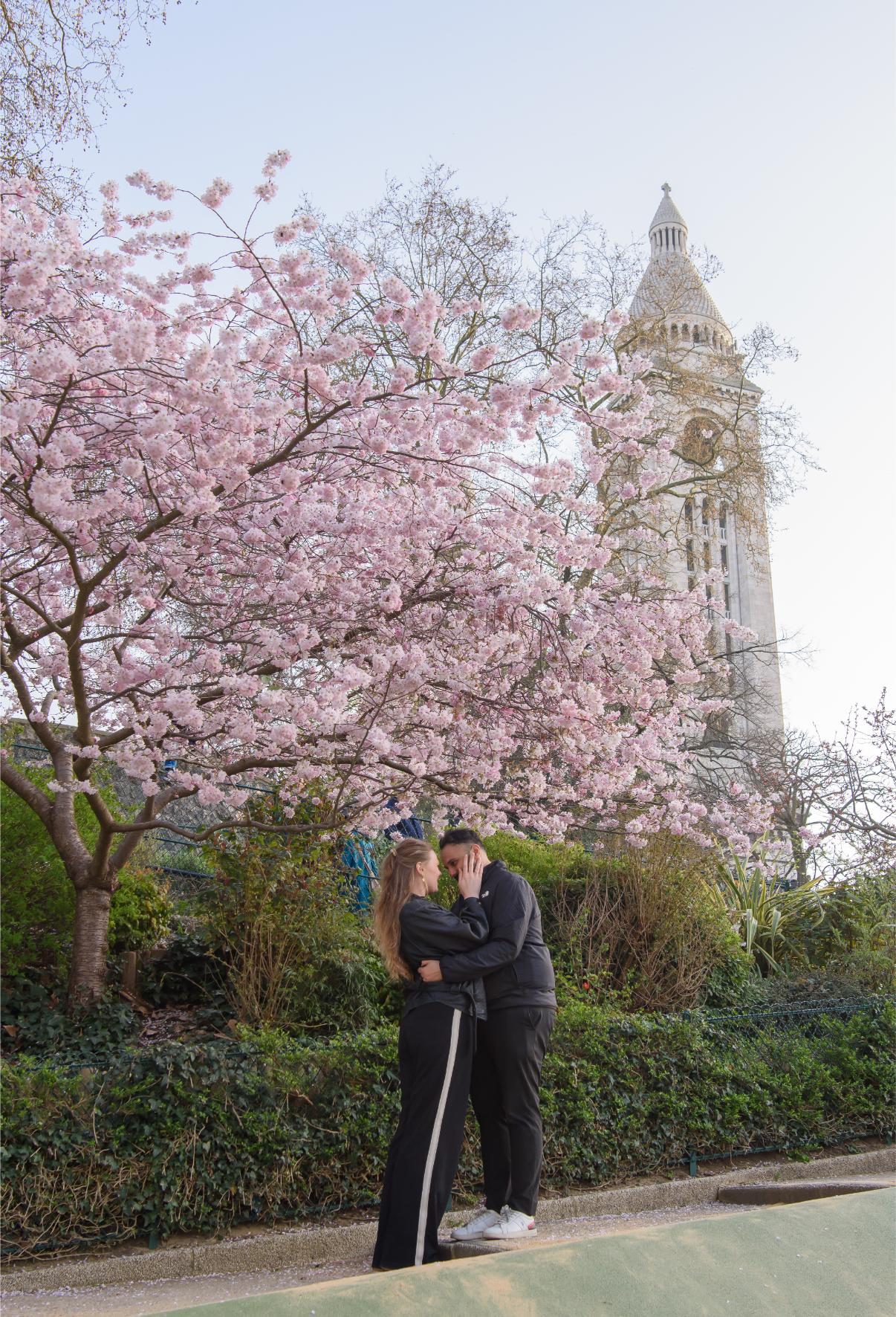 Couple sharing an intimate moment under a cherry blossom tree in Montmartre, with the Sacré-Coeur basilica tower visible in the background