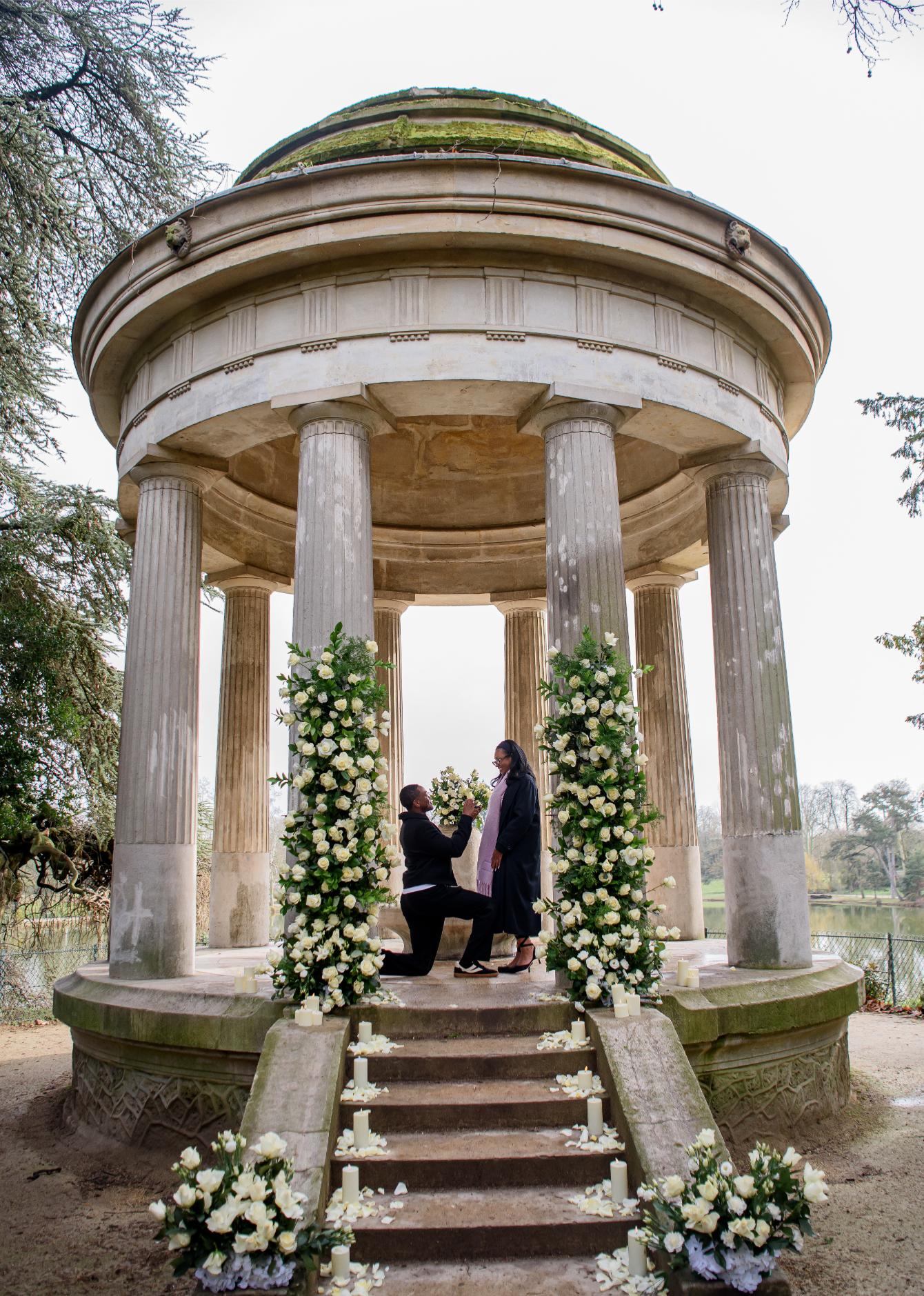 Man on one knee proposing inside the Parc Monceau rotunda in Paris, surrounded by white rose columns and candles
