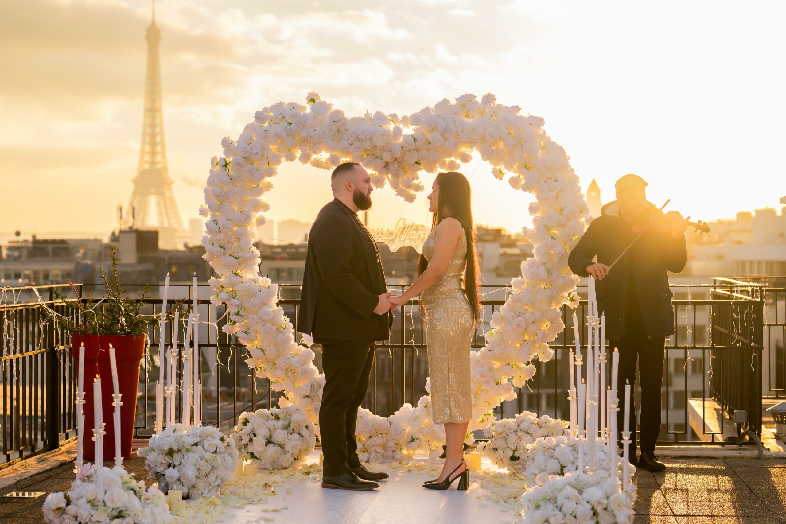 Couple standing under a white floral heart arch on a Paris rooftop terrace at sunset, with the Eiffel Tower in the background and a violinist playing nearby