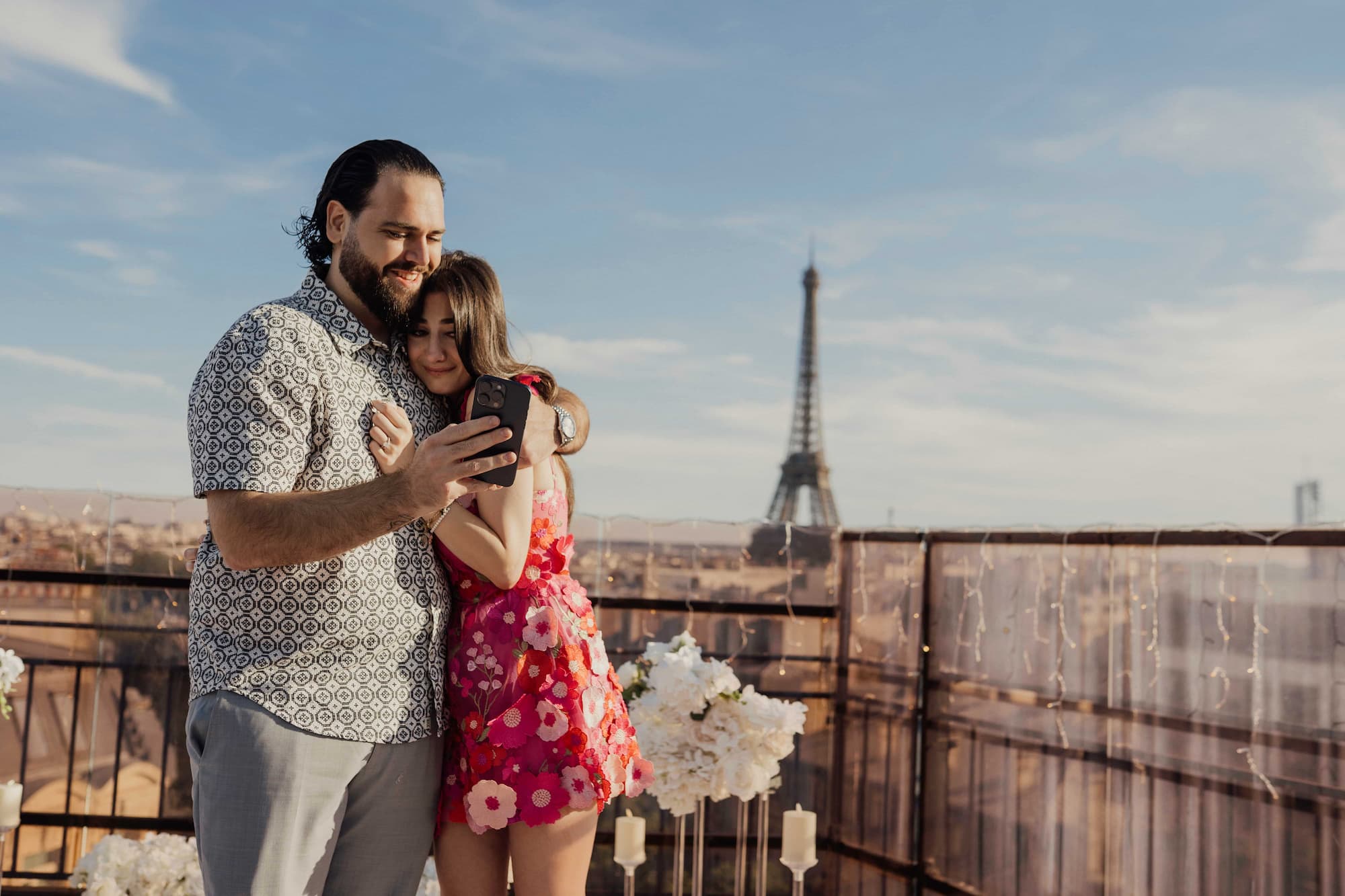 Newly engaged couple sharing a phone photo on a sunny Paris rooftop with Eiffel Tower view and floral décor
