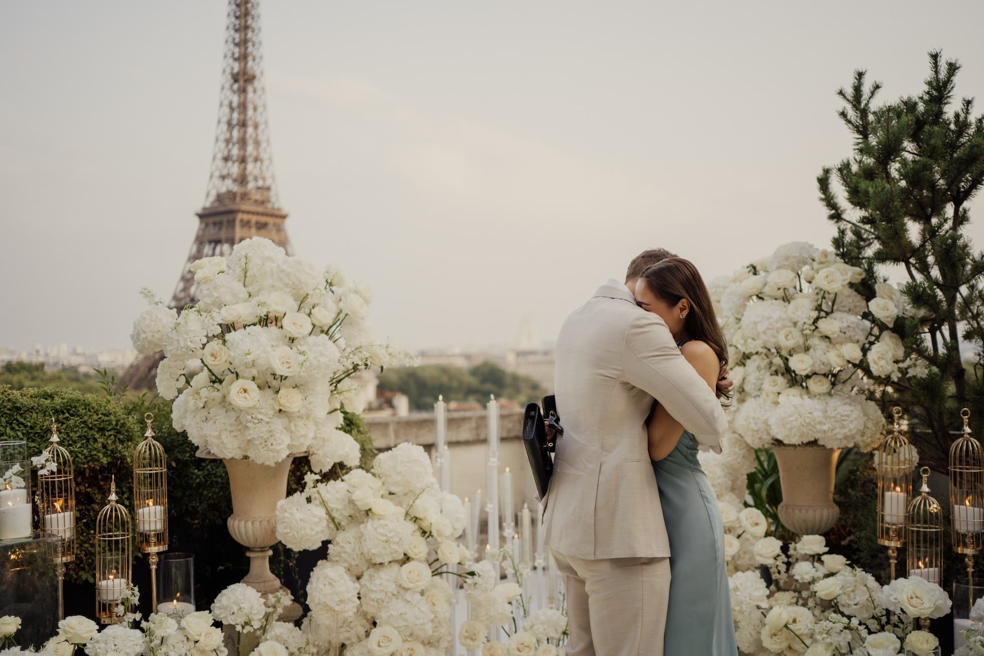 Couple embracing at a private rooftop proposal in Paris with white floral décor, candles and Eiffel Tower view