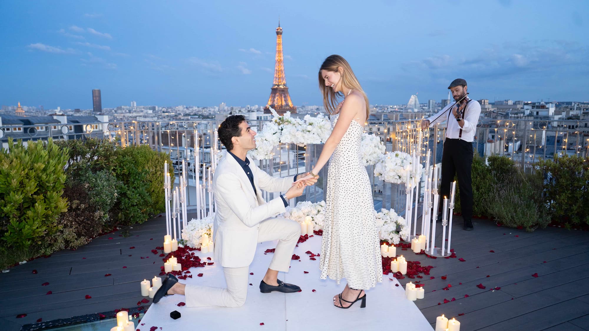 Man kneeling to propose on a Paris rooftop at dusk with live violinist, candles, rose petals and illuminated Eiffel Tower