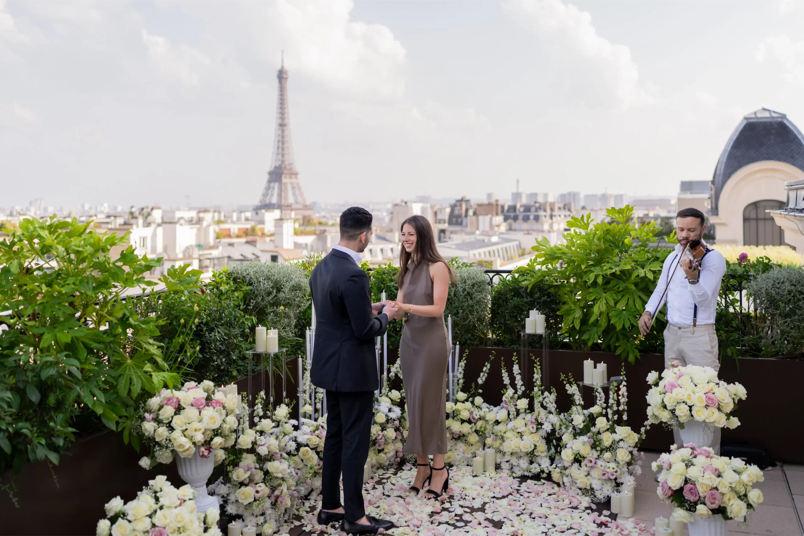 Rooftop marriage proposal in Paris with Eiffel Tower view, couple surrounded by white and pink roses and candles while a violinist plays