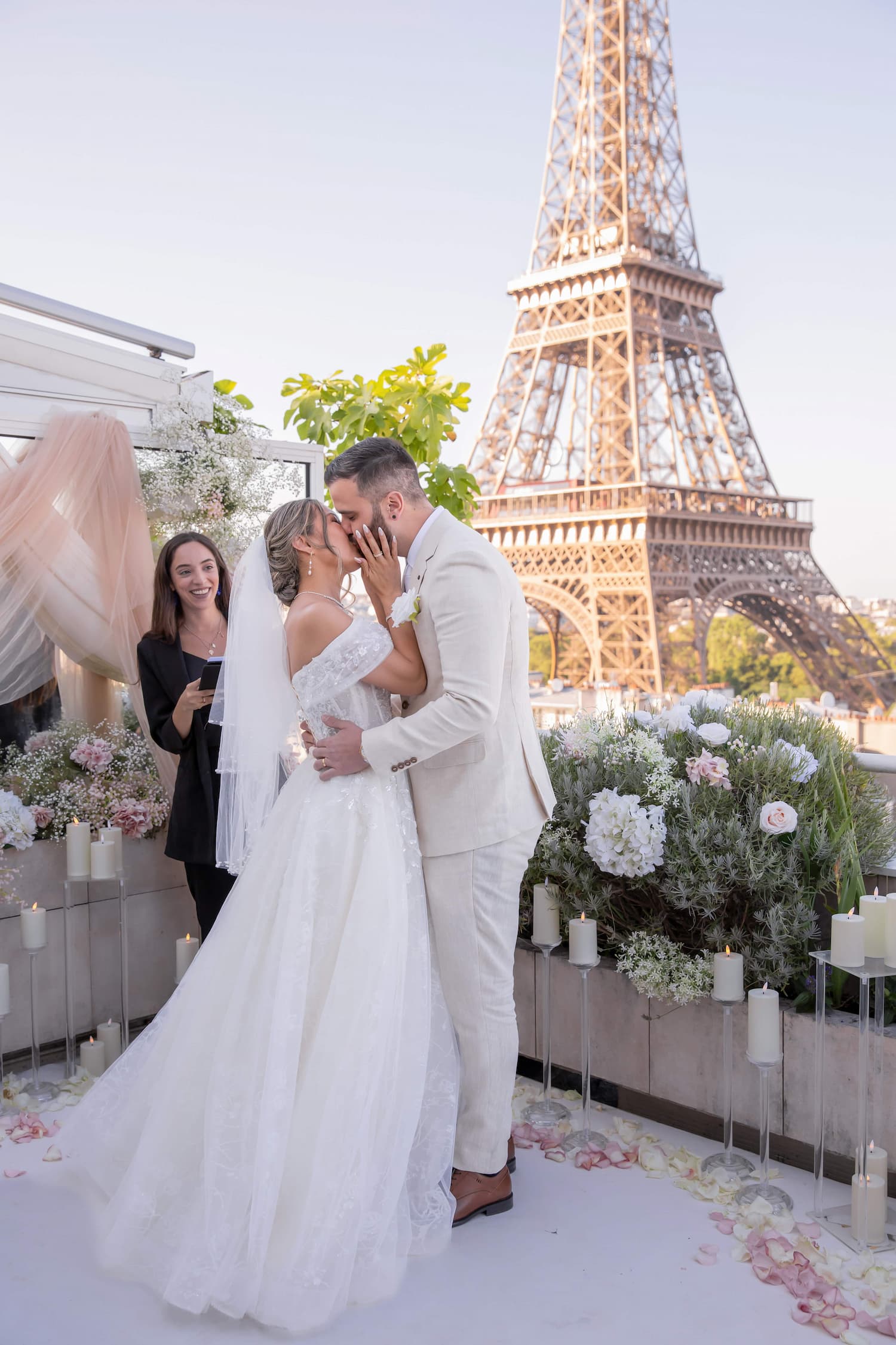 Bride and groom sharing their first kiss on a Paris rooftop terrace at dusk, with the Eiffel Tower directly behind them and a smiling officiant beside them