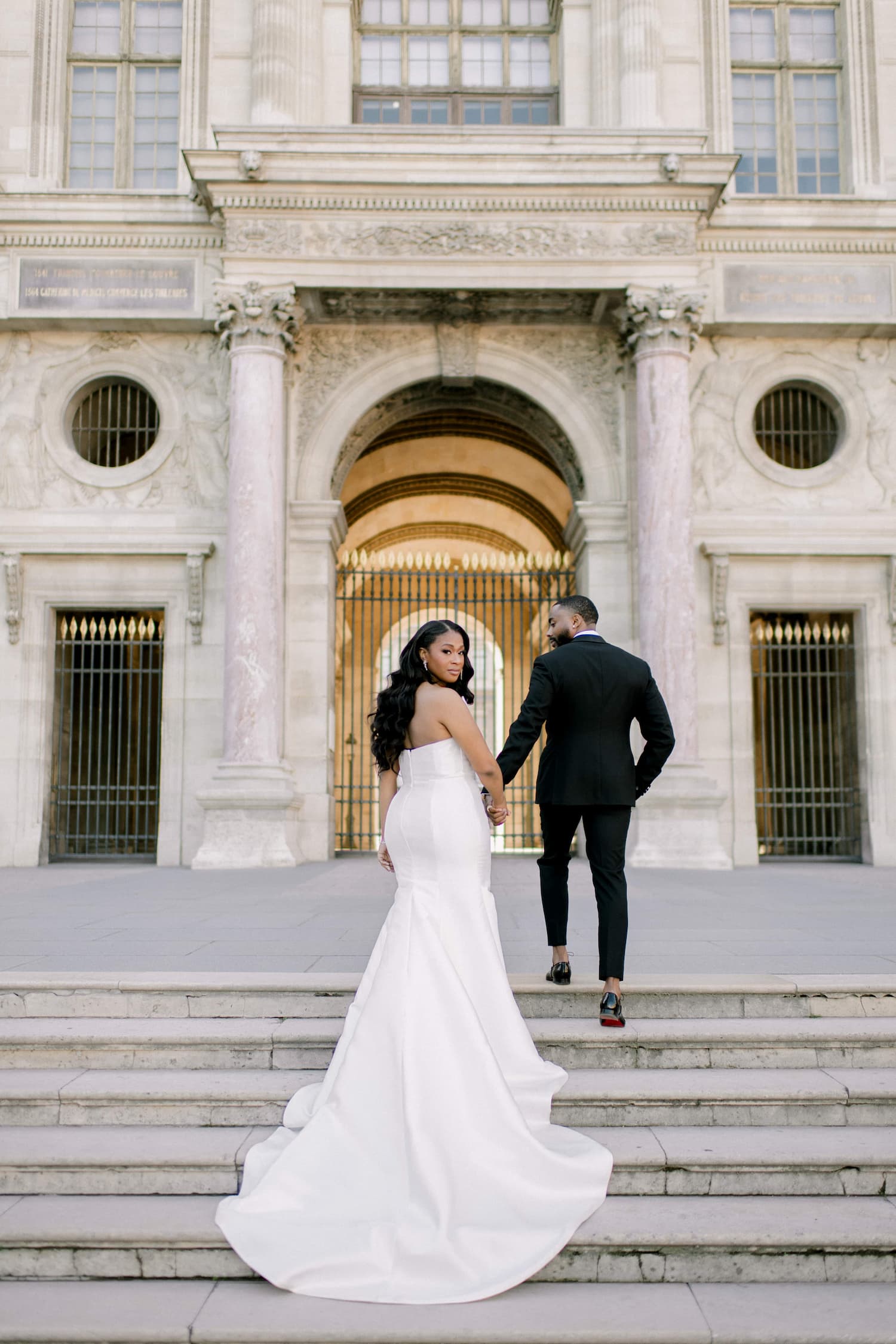 Bride in a white mermaid gown looking back over her shoulder while walking up the Louvre steps hand in hand with her groom during their Paris elopement shoot