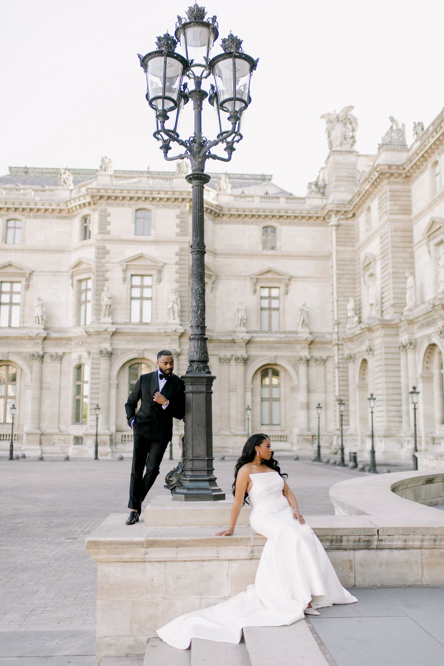 Bride in a white gown and groom in a black tuxedo posing by an ornate lamppost in the Louvre courtyard during their Paris elopement photo shoot
