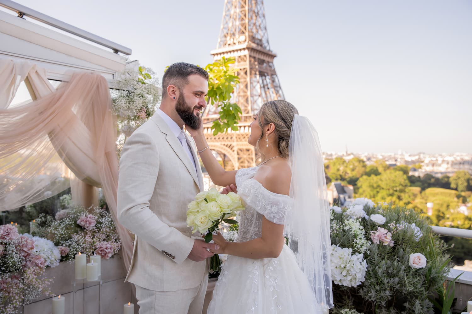 Bride and groom sharing an intimate moment on a Paris rooftop terrace surrounded by pink and white flowers, with the Eiffel Tower in the background at golden hour