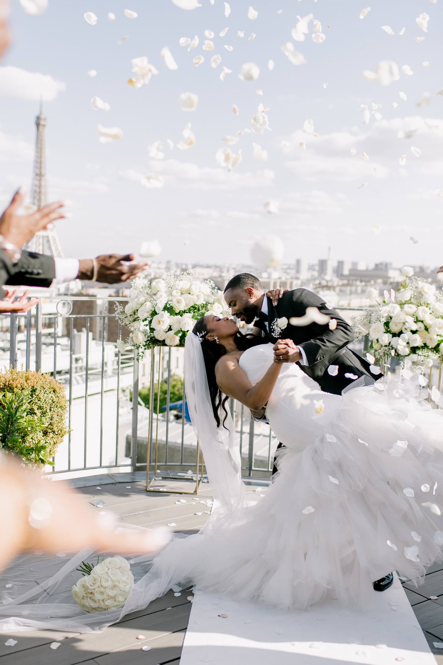 Groom dipping bride for a kiss on a Paris rooftop as white rose petals are tossed in the air, with the Eiffel Tower and city skyline in the background